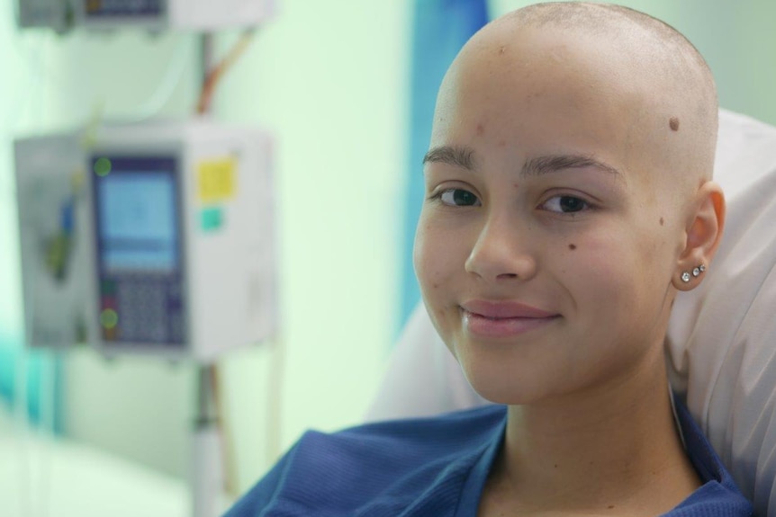 Sienna Martucci sitting up in a hospital bed smiling to the camera, with a hospital monitor in the background.