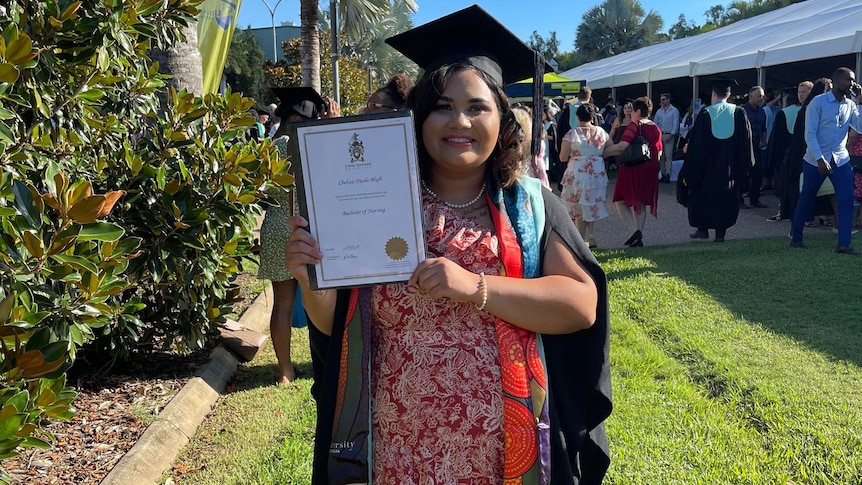 Chelsea holding her graduation certificate smiling, wearing graduation cap and gown, graduates behind her.