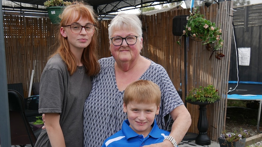 A woman stands with her arms around a 17-year-old girl and a younger boy, standing in a backyard