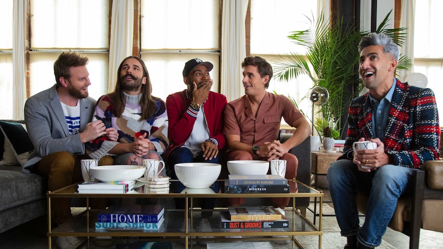 The cast stand shoulder to shoulder in a kitchen, smiling