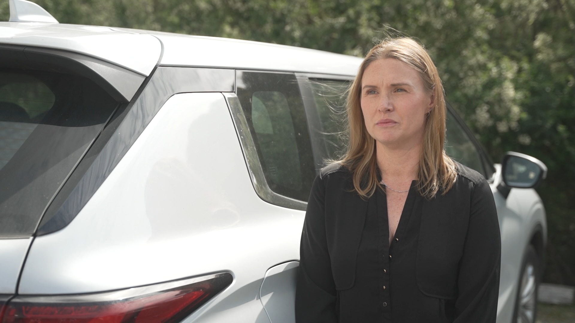 A woman with brown shoulder-length hair stands beside her car.