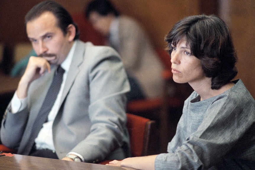 A woman with short brown hair and a man in a grey suit sit in a hearing room.