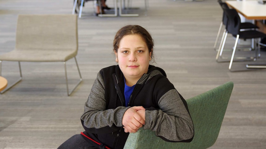 A young teenager sits in the middle of a quiet library looking directly at the camera
