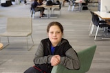 A young teenager sits in the middle of a quiet library looking directly at the camera