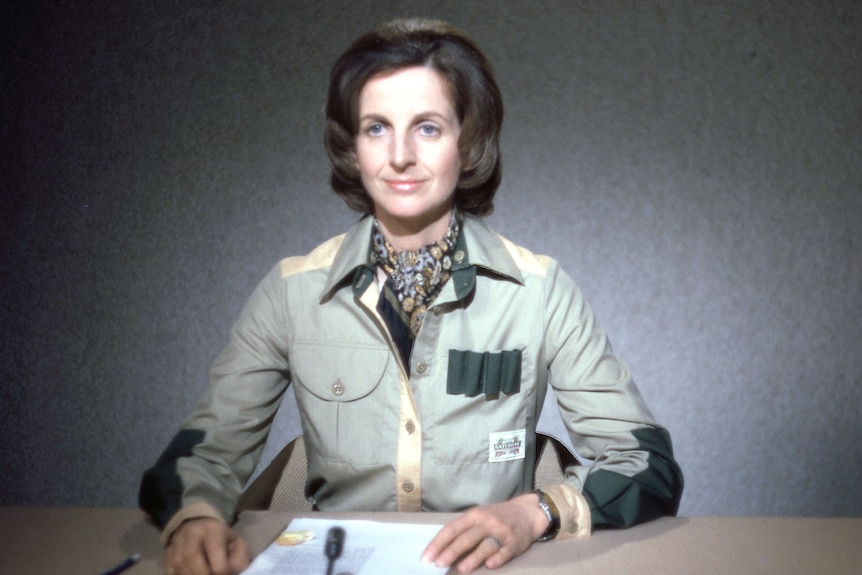 A woman with short bob and wearing a green safari suit sits at a newsreader desk