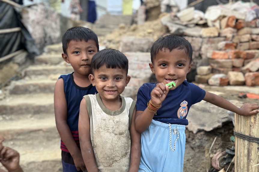 Three young Rohingya boys stand near bricks and stairs, smiling