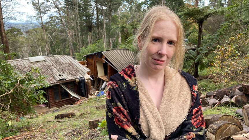Kate Naughton stands, arms folded, in front of her rubble-strewn home nestled among tall trees at Ferny Creek.