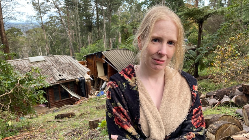 Kate Naughton stands, arms folded, in front of her rubble-strewn home nestled among tall trees at Ferny Creek.