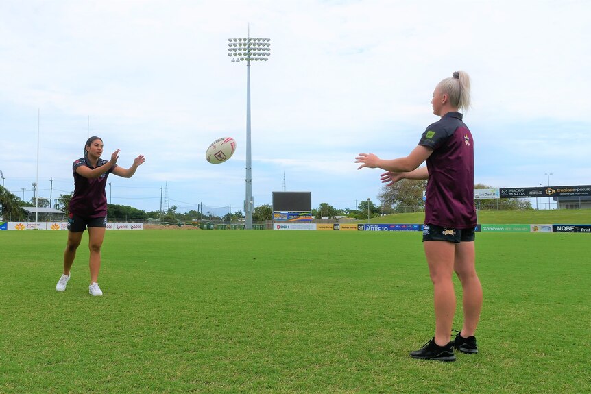 Two female footy players in maroon shirts passing a ball on the field.