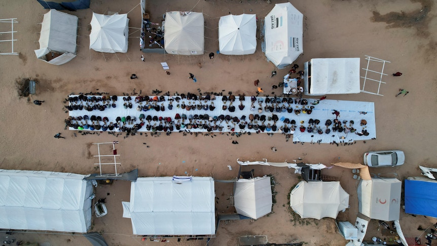 A drone image of about a hundred people sitting down on a long white mat to eat, with relief tents around them.