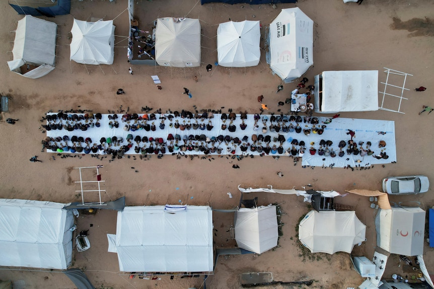 A drone image of about a hundred people sitting down on a long white mat to eat, with relief tents around them.