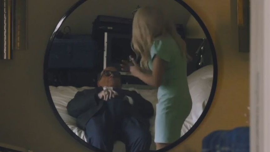 Rudy Giuliani lies down on a bed with his hand in his pants next to the actor Maria Bakalova