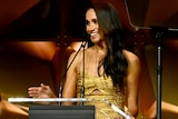Meghan, Duchess of Sussex, wears a gold strapless dress and speaks into a microphone on stage.