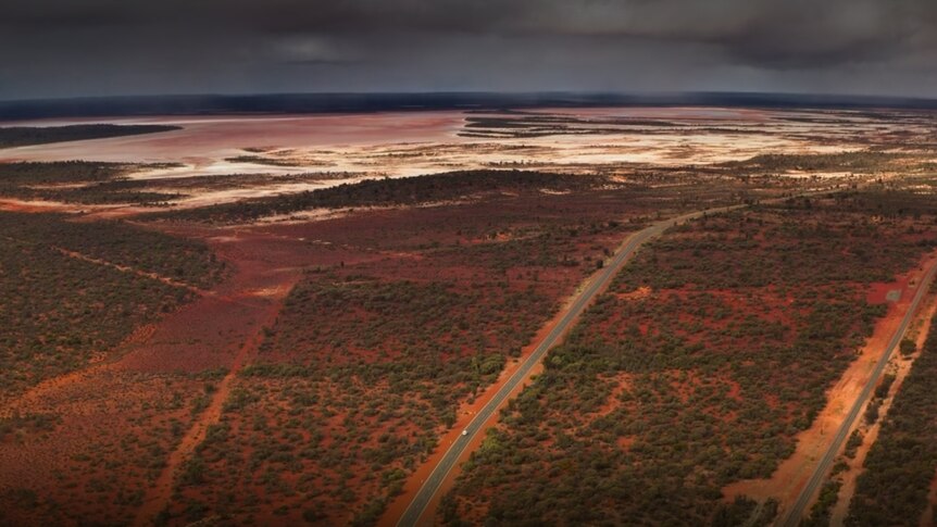 A small white car drives on a road through the desert of Australia