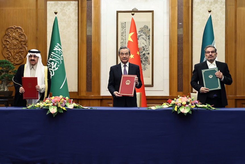 Three men stand behind table holding up folders for cameras, flags of Saudia Arabia, China, Iran in background.