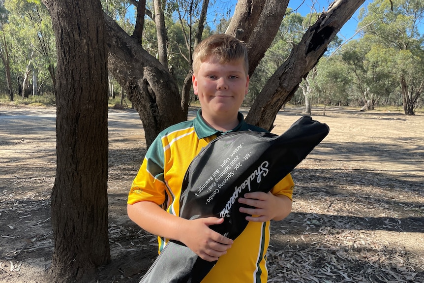 Zach is smiling holding his fishing rod kit wearing a school uniform near his home in Echuca