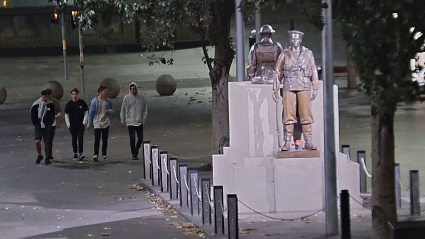Five young men in casual clothing walk towards a cenotaph statue of two soldiers.