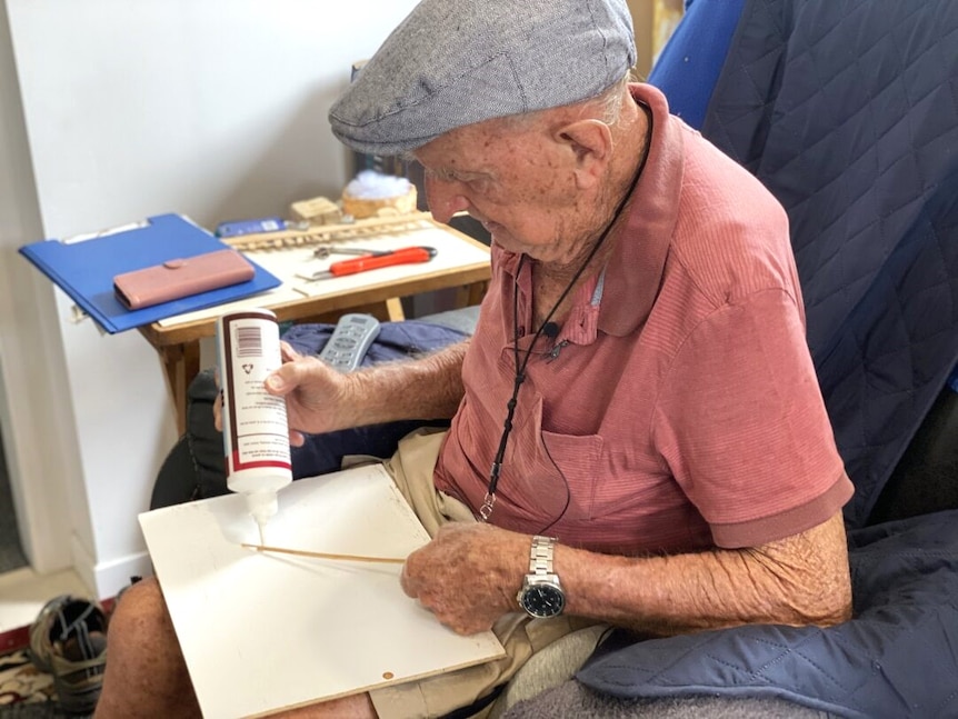An older man sitting in a chair holding a piece of wood and a tube of glue.