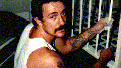 An old coloured photo of moustached man with tattoos on arm, a white vest, painting a window bar white, looks up at camera.