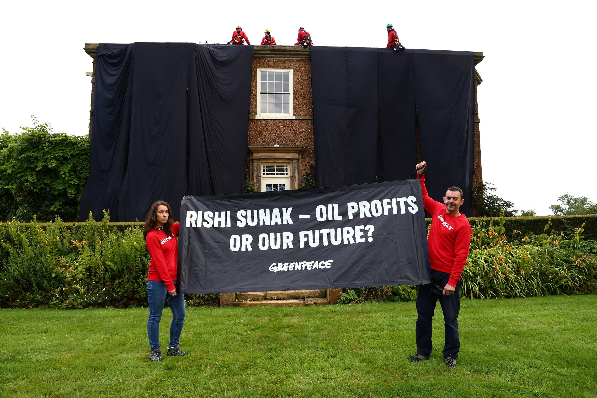 Two people wearing red holding a black banner with text and a two storey house draped in black cloth behind them