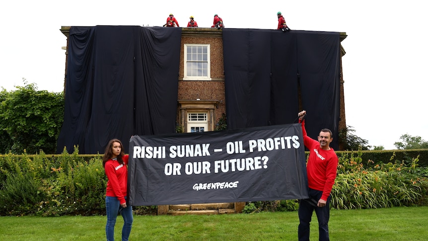 Two people wearing red holding a black banner with text and a two storey house draped in black cloth behind them