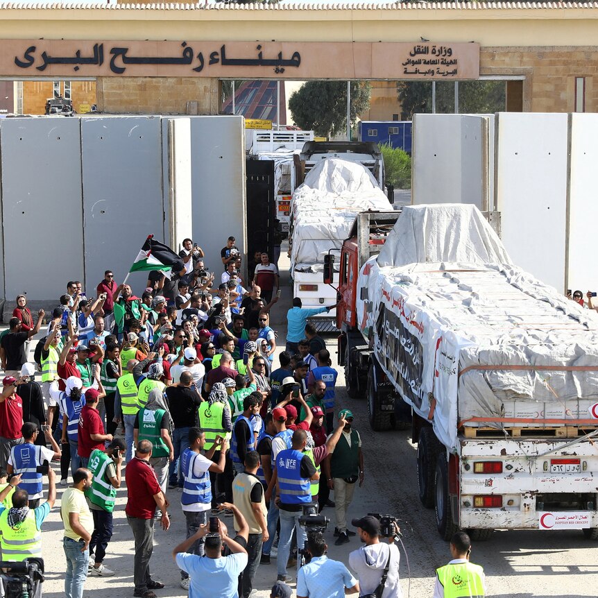 A crowd of people, some wearing hi-vis vests, gather beside several trucks loaded with covered goods as they emerge from a wall.