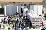 A crowd of people, some wearing hi-vis vests, gather beside several trucks loaded with covered goods as they emerge from a wall.