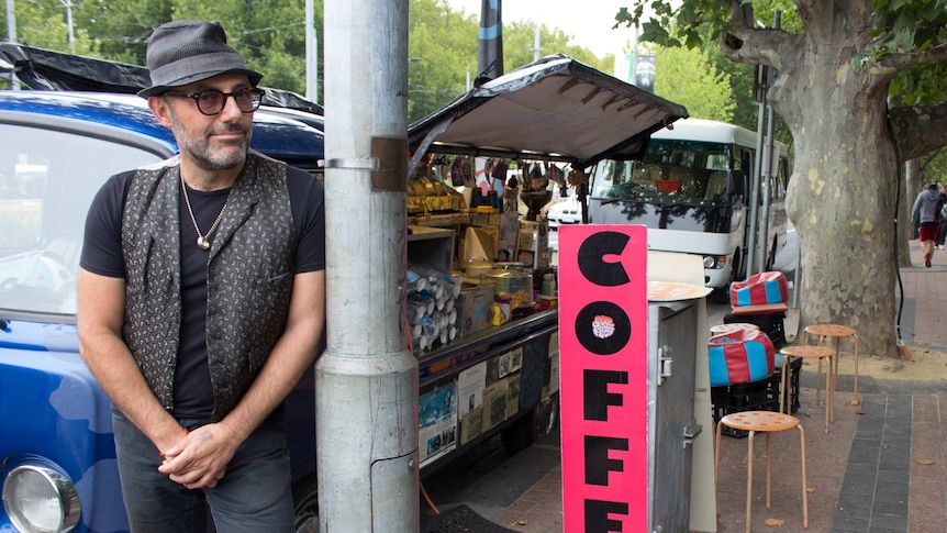 A man stands next to a coffee van, seats on footpath.