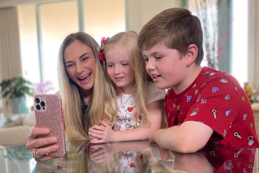 A mother and her young daughter and son sit at a dining table looking at a smartphone and smiling.