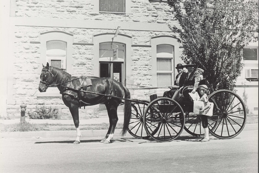 A horse and cart collecting a newspaper from a child at the front of a building.