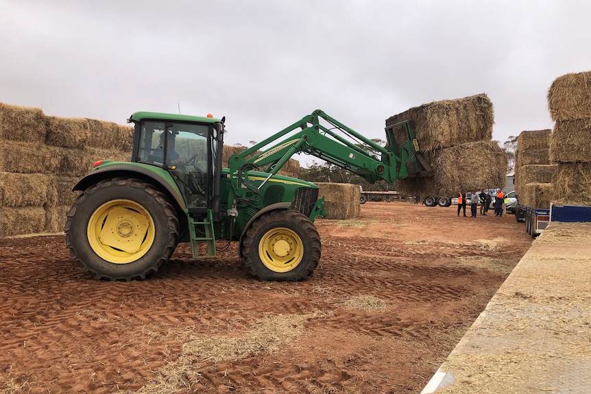 A green tractor with two bales of hay on its forks, travelling from left to right across the frame 