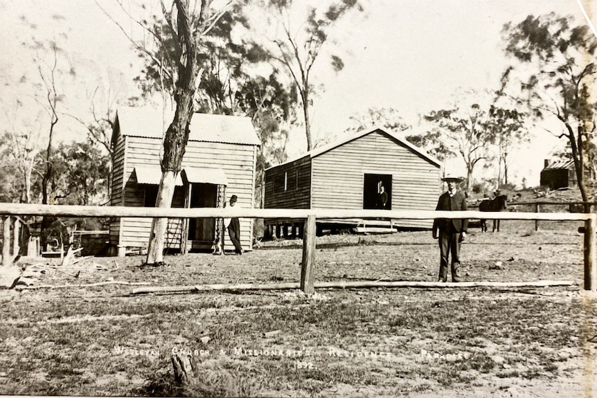 black and white image of two wooden buildings, with a man standing in a suit in the foreground