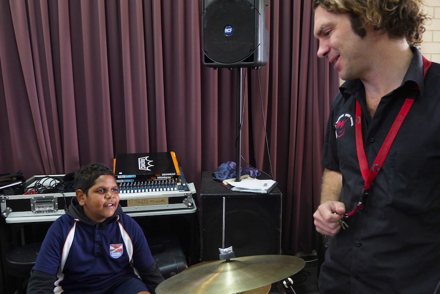 Man with curly short hair wearing black shirt standing next to drum kit speaking to boy behind the kit.