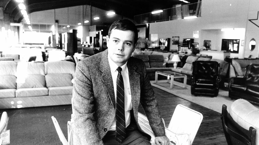 Black and white photograph of a young man in a tweed jacket sitting in the middle of a furniture store.