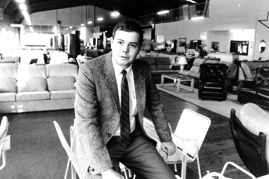Black and white photograph of a young man in a tweed jacket sitting in the middle of a furniture store.