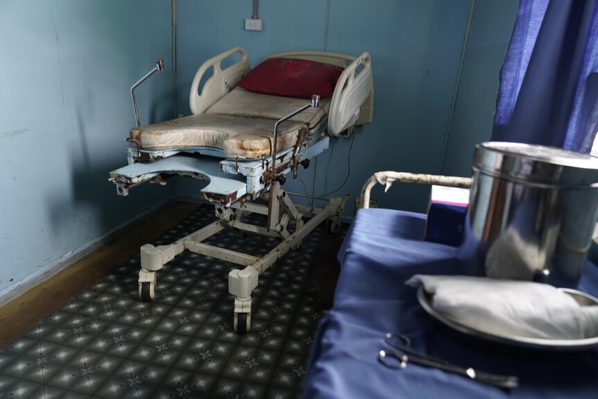 A dirty looking hospital bed sits in the corner of a room. A table with stainless steel equipment is also in frame