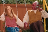A 30-something woman and a masked man in 70s dress hold hands while smiling broadly and swinging on a yellow backyard swing set.