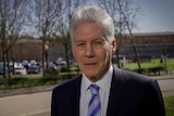 A man with white hair and a suit looks at the camera. In the background are the brick walls of the prison