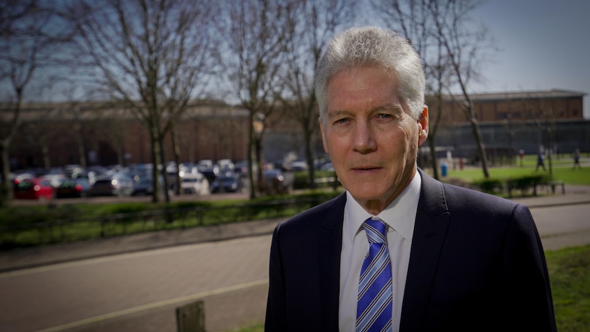 A man with white hair and a suit looks at the camera. In the background are the brick walls of the prison