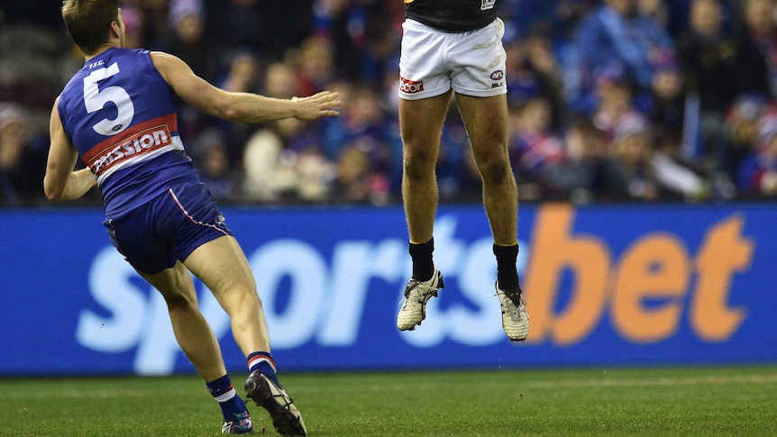 Two AFL players in the foreground with a betting company sign blurred in the background