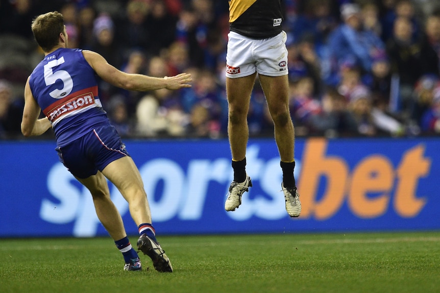 Two AFL players in the foreground with a betting company sign blurred in the background