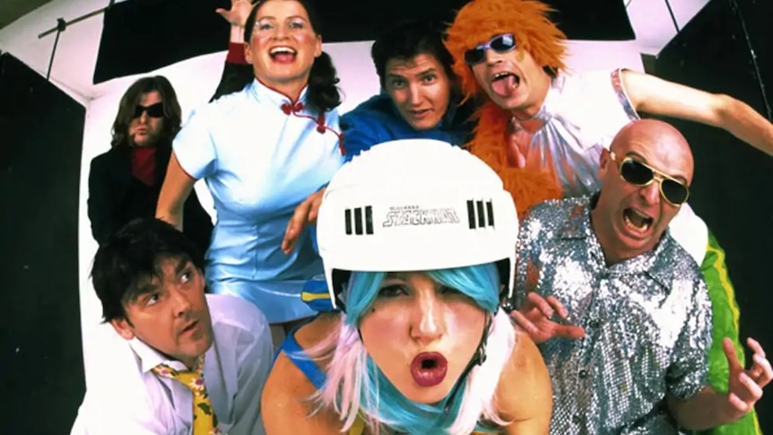 A fish-eye style photo of seven people wearing costumes like bike helmets, sequined shirts, and a cheongsam.