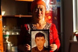 A woman wearing an orange headwrap and red top looks into the camera as she holds up a photograph of a man