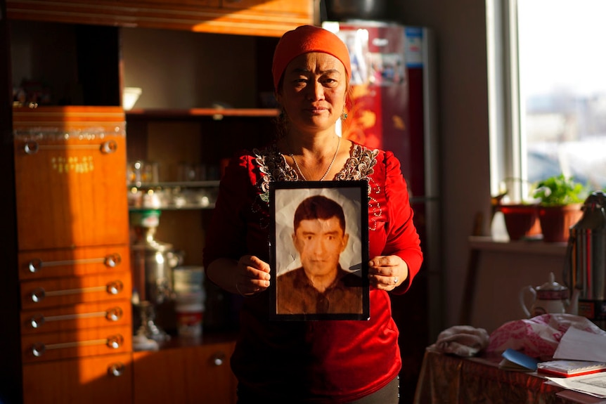 A woman wearing an orange headwrap and red top looks into the camera as she holds up a photograph of a man