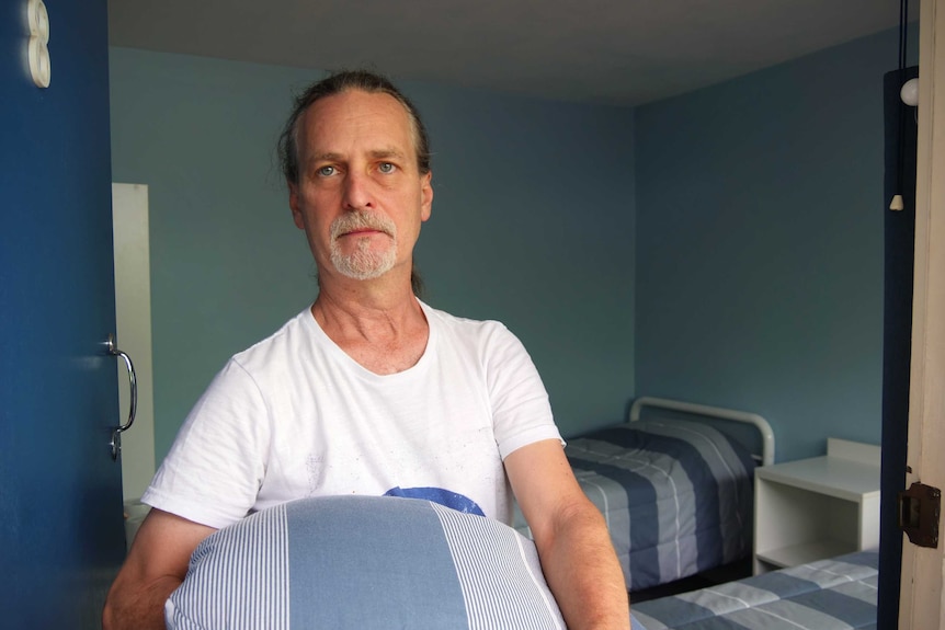 A middle-aged man with long hair and a goatee stands in a hostel room holding bedding.