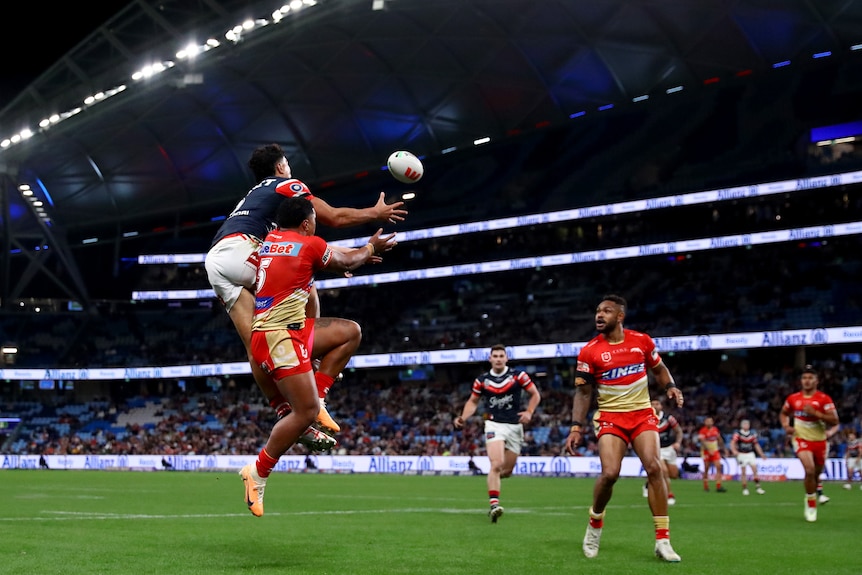 A Roosters NRL player reaches out with his arms to collect the ball as he flies through the air ahead of a Dolphins defender.