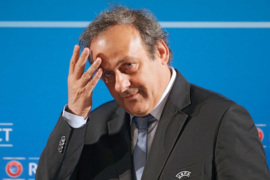 Michel Platini, wearing a suit, touches his forehead and bows his head slightly.