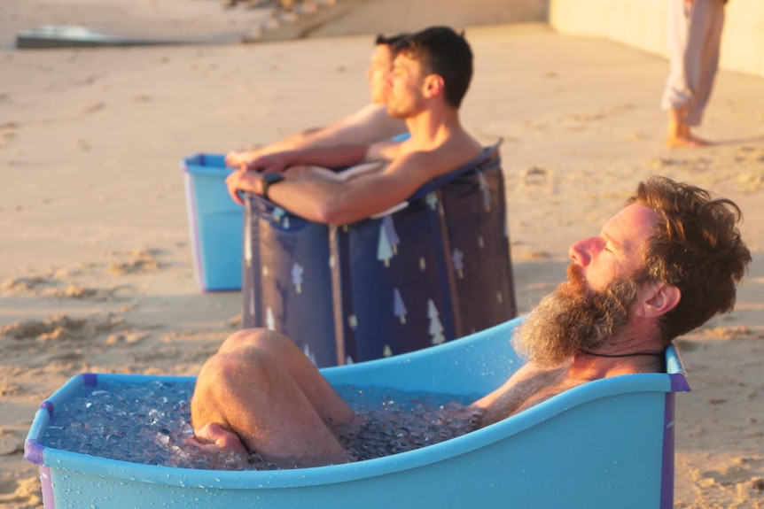 Trois hommes sont assis dans de petits bains bleus séparés remplis de glace, les yeux fermés 