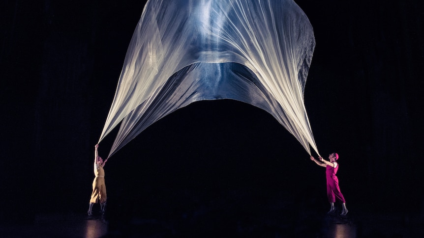 A woman in yellow and a man in pink hold up a large sheer fabric filled with air with light inside it
