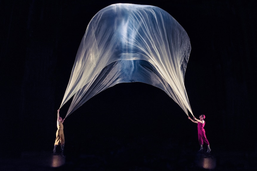 A woman in yellow and a man in pink hold up a large sheer fabric filled with air with light inside it